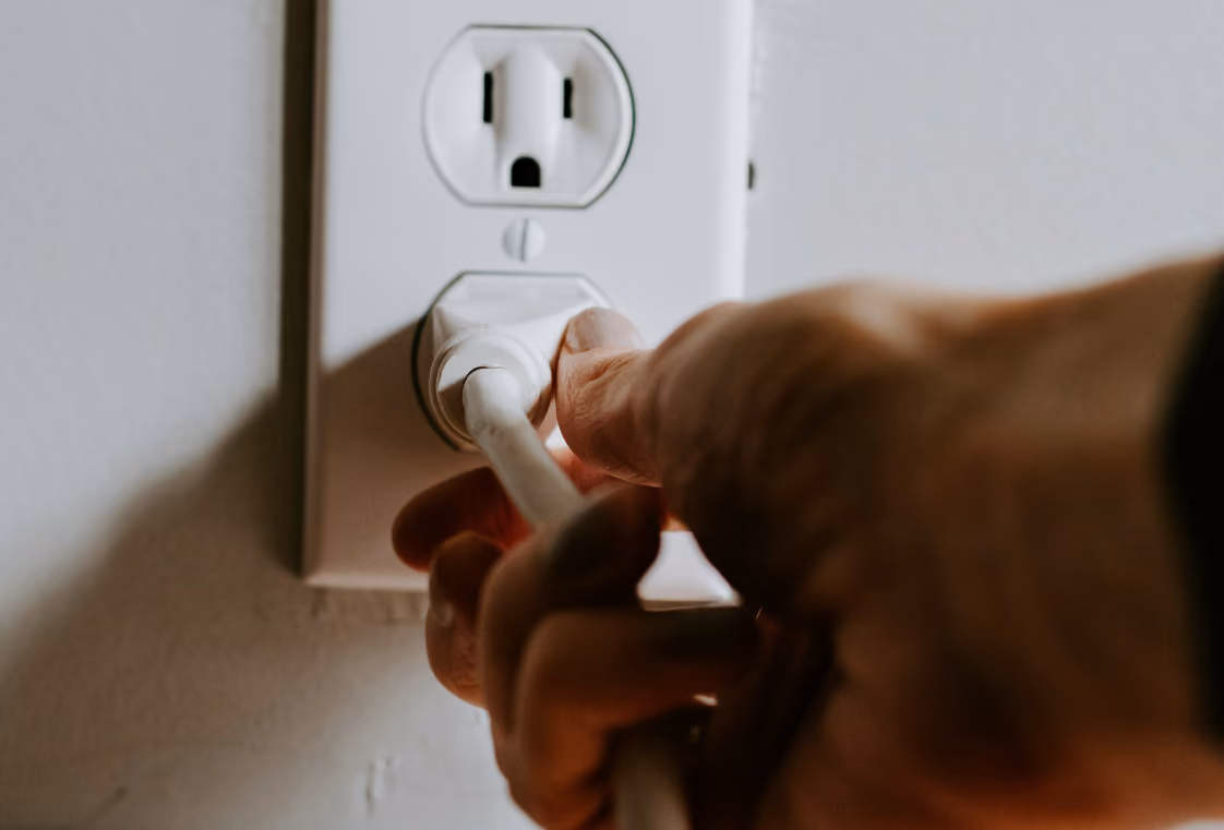 Hand plugging a white cord into a duplex wall outlet on a light-colored wall