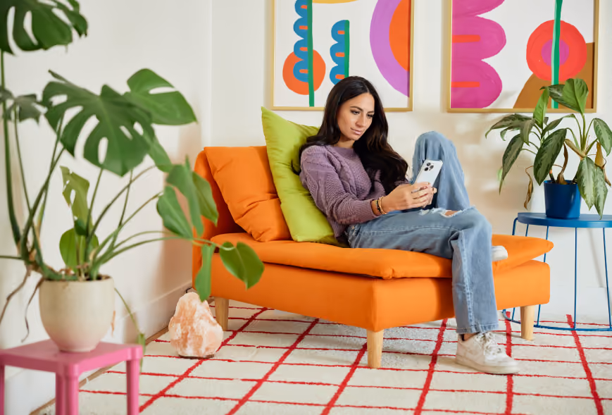 Woman on a vivid orange chaise using a phone beside leafy plants and bold abstract art