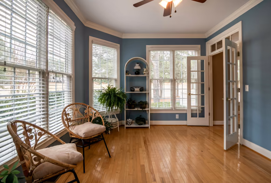 Sunlit sitting room with rattan chairs, plants, white blinds, and French doors on hardwood floors