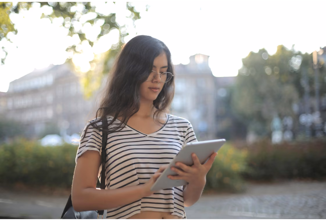 Woman in glasses using a tablet outdoors with blurred trees and city buildings behind her