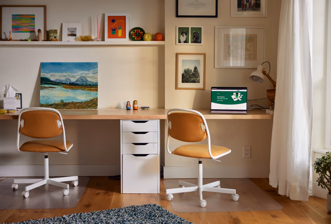Shared home office with a long wood desk, two orange chairs, art, and a laptop open on the desk