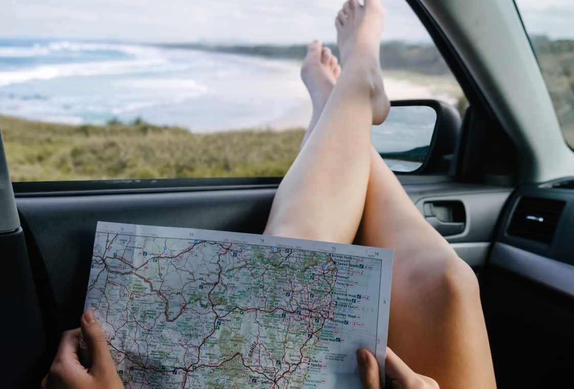 Passenger holding a coastal road map with legs on the dash and a sandy beach visible outside