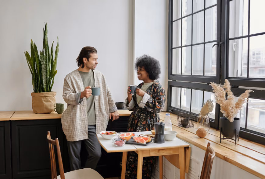 Two people in robes chat over breakfast at a small table in a bright kitchen beside a large black-framed window