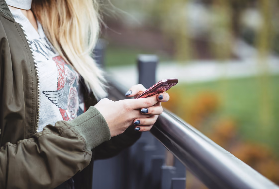 Side view of a person in an olive bomber jacket using a rose-gold smartphone while leaning on a silver railing outdoors