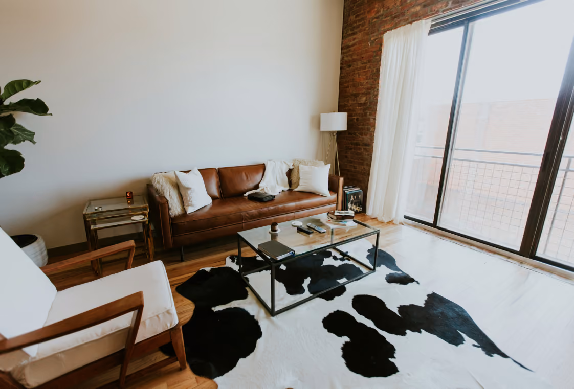 Airy living room with brown leather sofa, glass coffee table on a cowhide rug, exposed brick, and balcony doors
