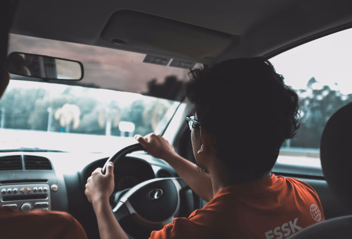 Back-seat view of a driver in glasses and a red shirt steering along a road past blurred trees