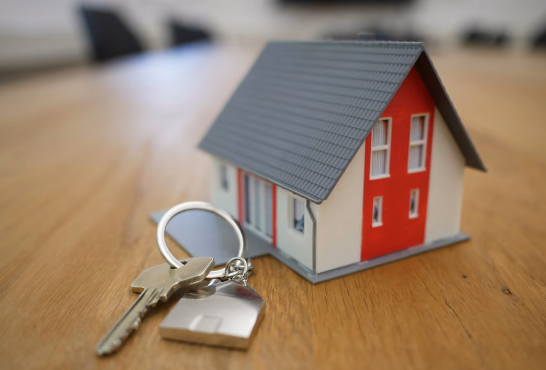 Small model house beside keys with a house-shaped metal fob on a light wood surface