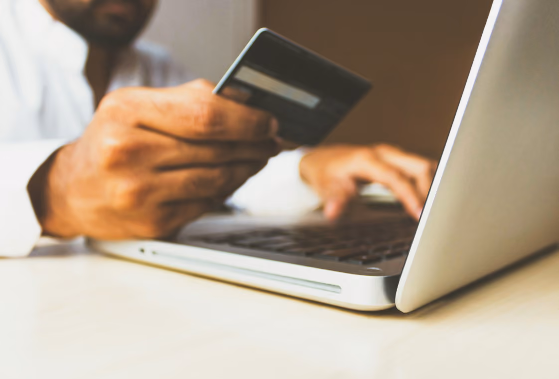Hands on a laptop keyboard while holding a credit card toward the screen on a wooden table