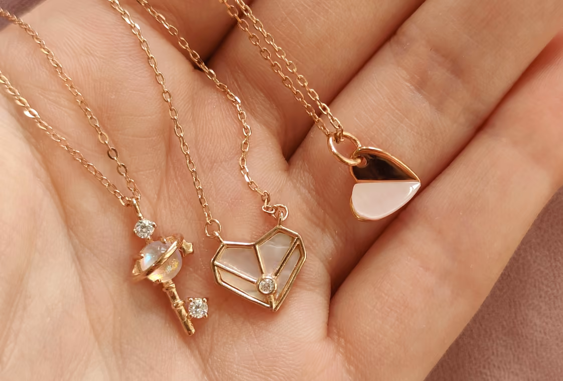 Three delicate rose gold pendant necklaces resting in an open palm over a dusty rose background