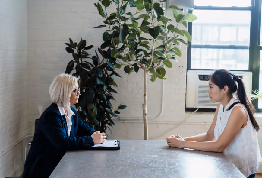 Two women seated across a conference table in a bright office, one with a clipboard, in a focused meeting