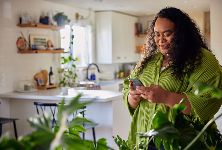 Smiling person in a lime green shirt using a smartphone in a bright kitchen with white cabinets and shelves