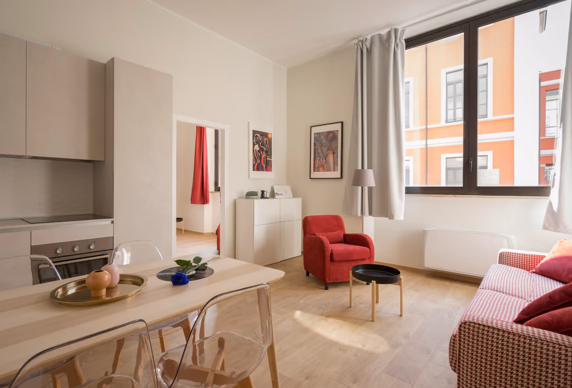 Sunlit open-plan apartment with wood dining set, sleek gray kitchen, and red accent seating by tall windows
