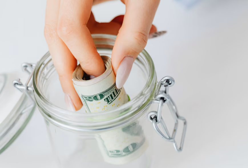 Hand lowering a roll of US bills into a clear swing-top glass jar on a white counter
