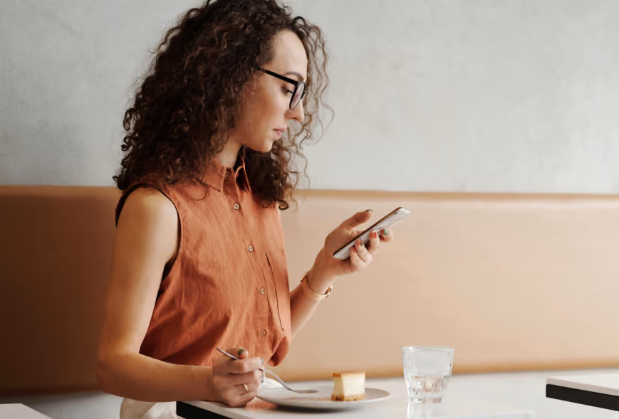 Woman with curly hair and glasses using her phone over cheesecake at a bright café table