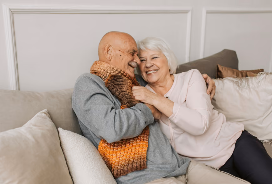 Senior couple laughing while cuddling on a sofa with neutral pillows and paneled wall behind them