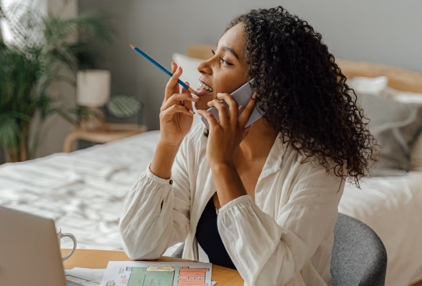 Smiling woman on a phone call at a desk with floor plans, laptop, and mug in a sunlit room
