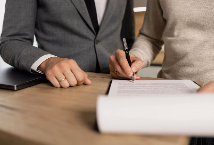 Two people signing paperwork on a clipboard at a light wood desk beside a closed laptop