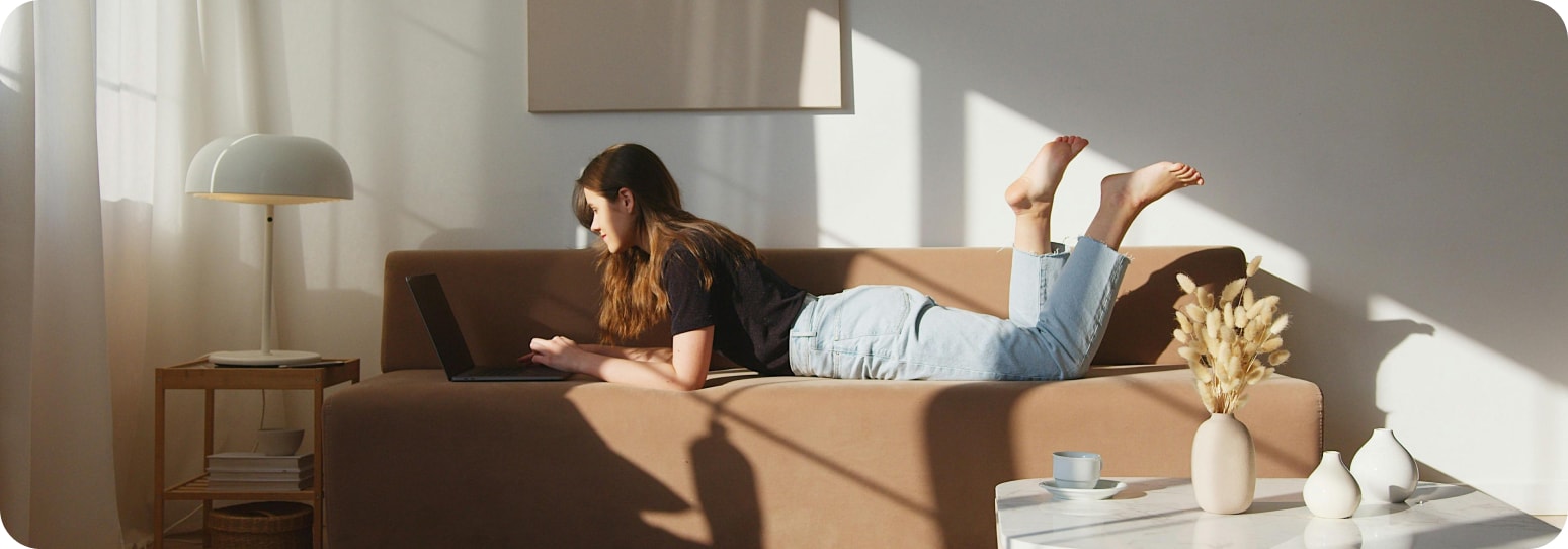 Woman lying on her stomach on her couch in an apartment looking at her laptop
