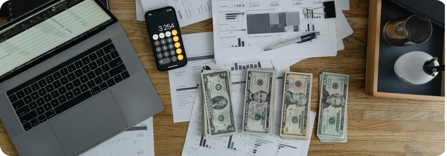 A busy looking desk with financial papers, a laptop, calculator, and money.