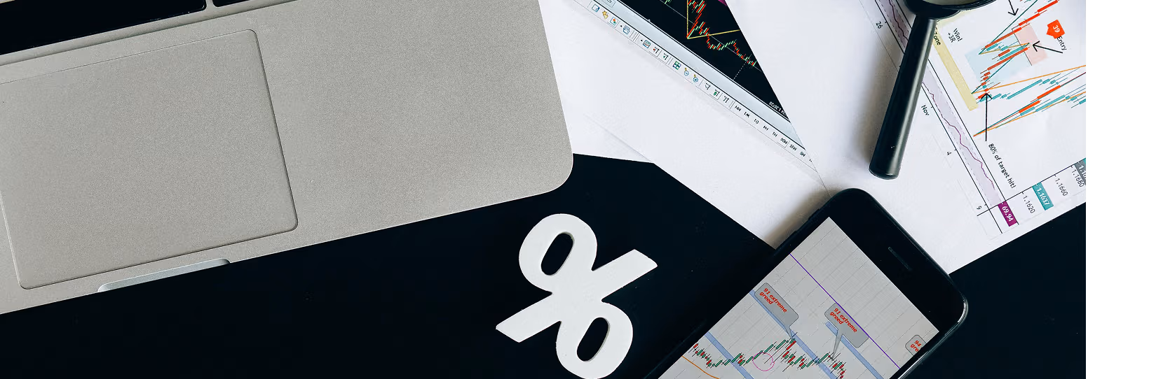 an birds eye view of a laptop, smart phone, a percentage sign, and papers with stock charts