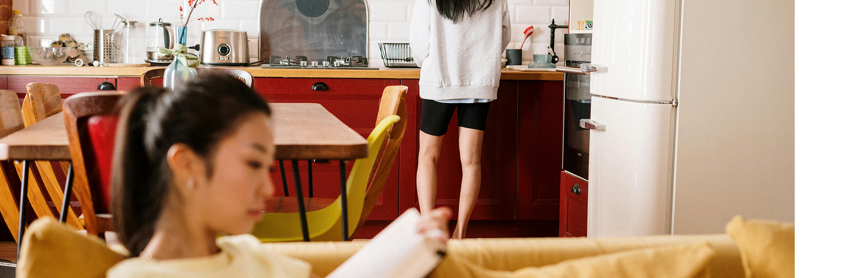 a person sitting on a couch in a living room laying on a couch, with another person in the background doing dishes in the kitchen