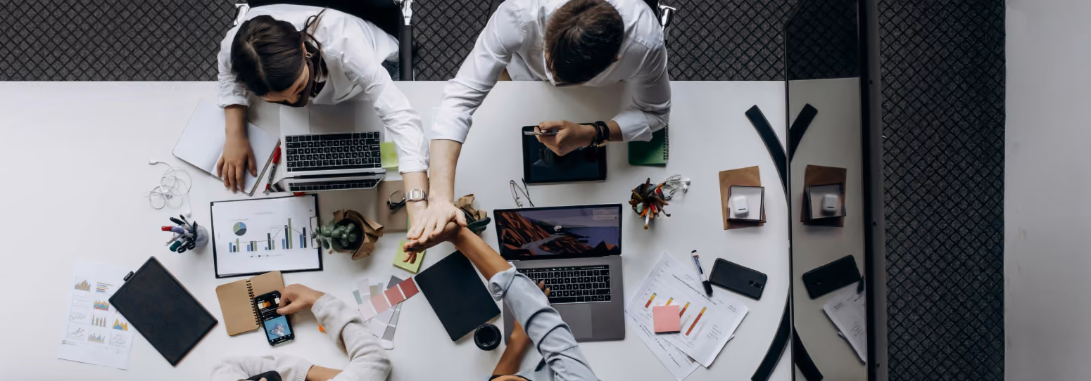 A group of 4 people dressen in business formal attire sitting at a meeting table putting their hands together