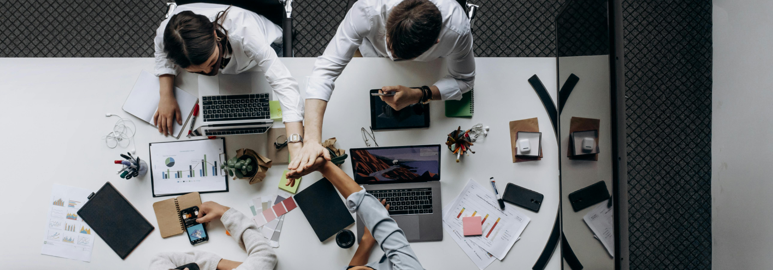 A group of 4 people dressen in business formal attire sitting at a meeting table putting their hands together