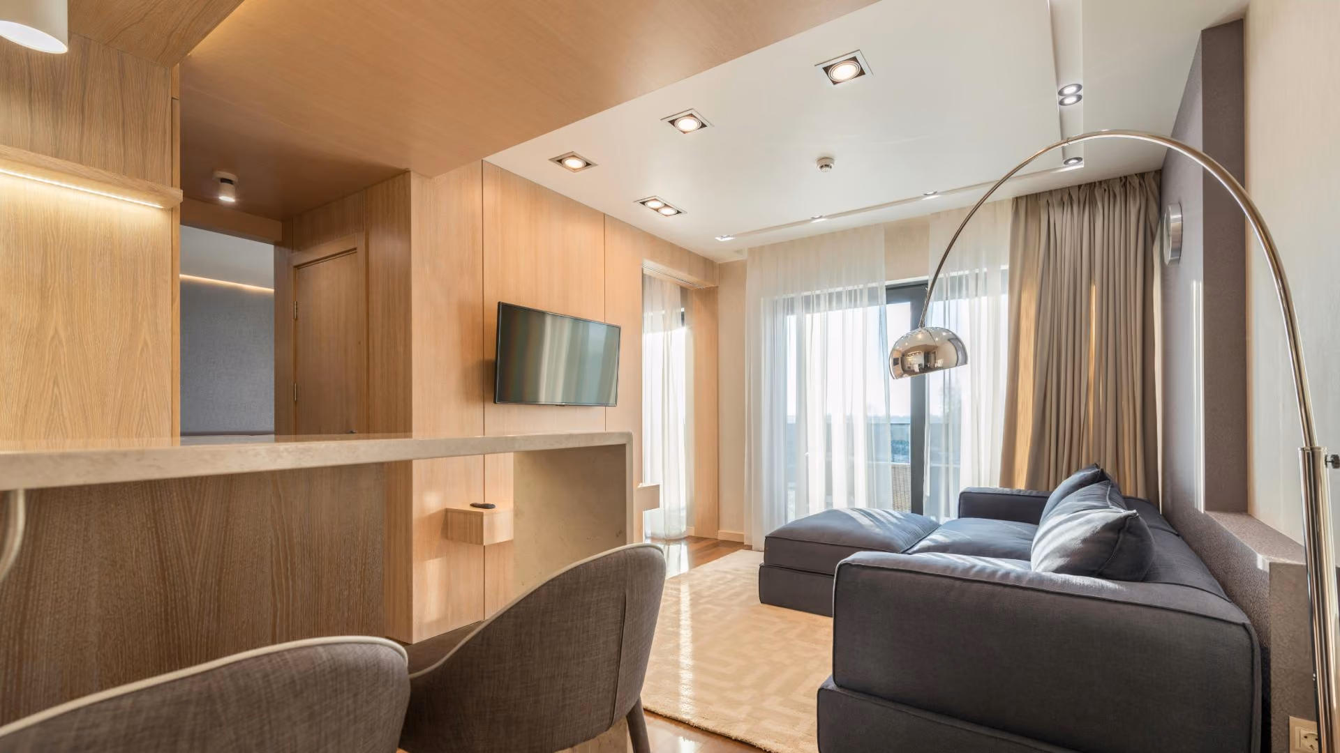 Modern apartment living room featuring a dark grey sectional sofa, light wood wall paneling, and a large arched chrome floor lamp.