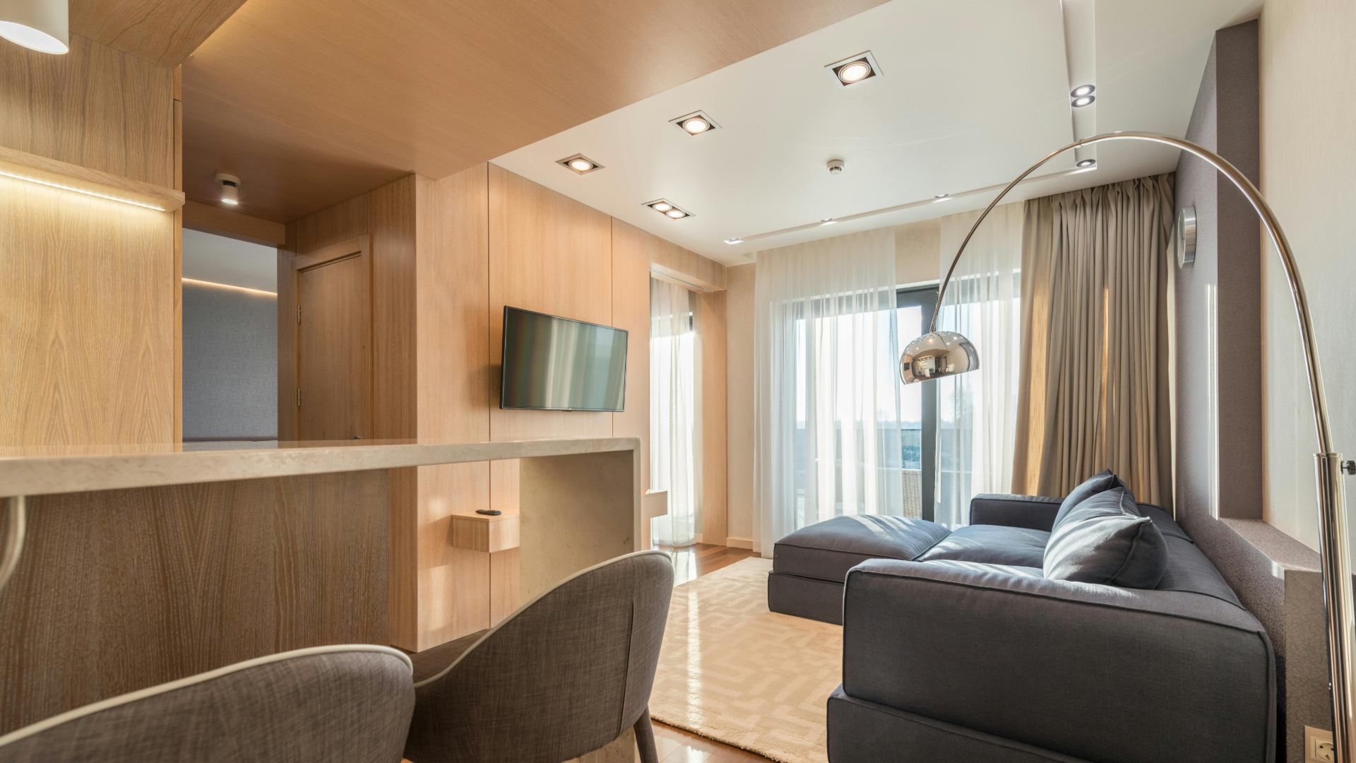 Modern apartment living room featuring a dark grey sectional sofa, light wood wall paneling, and a large arched chrome floor lamp.
