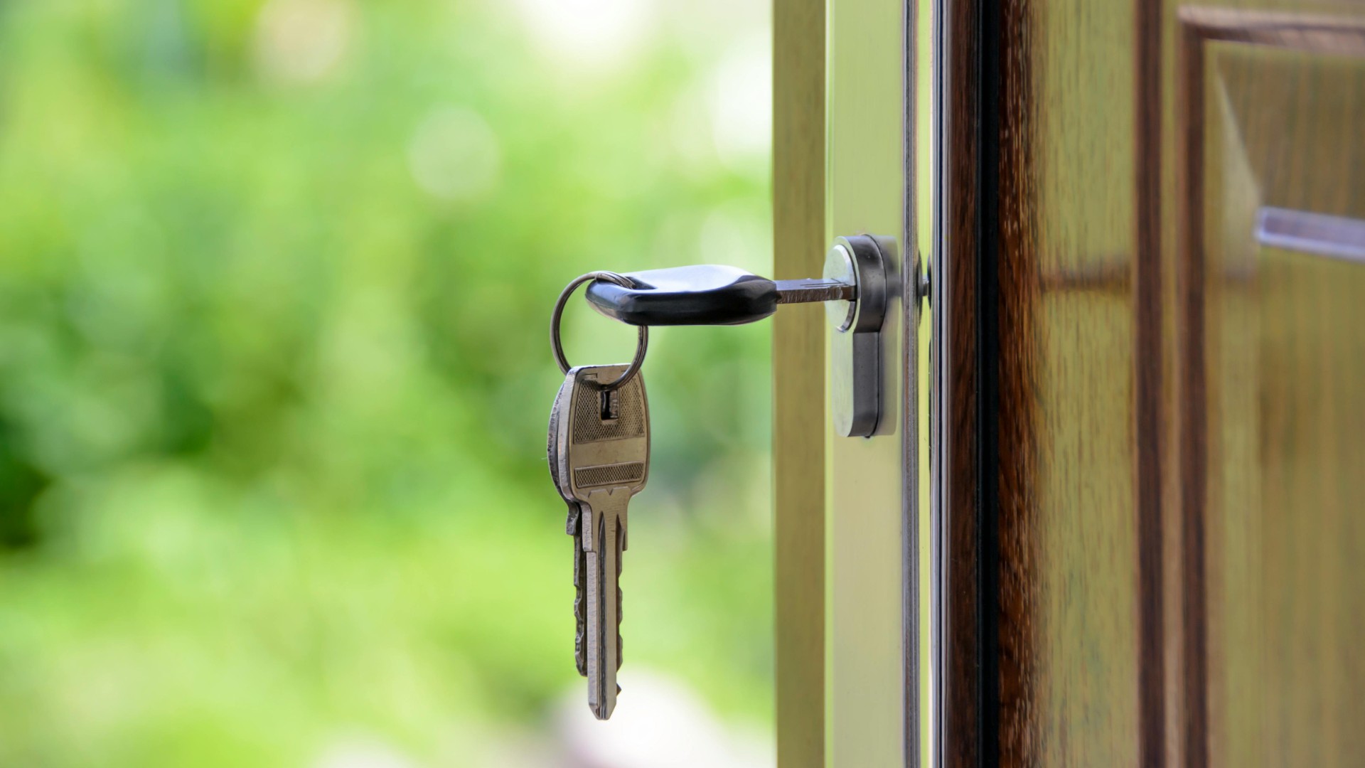 A set of silver keys hanging from a lock in an open wooden door, with a soft, blurred green outdoor background.