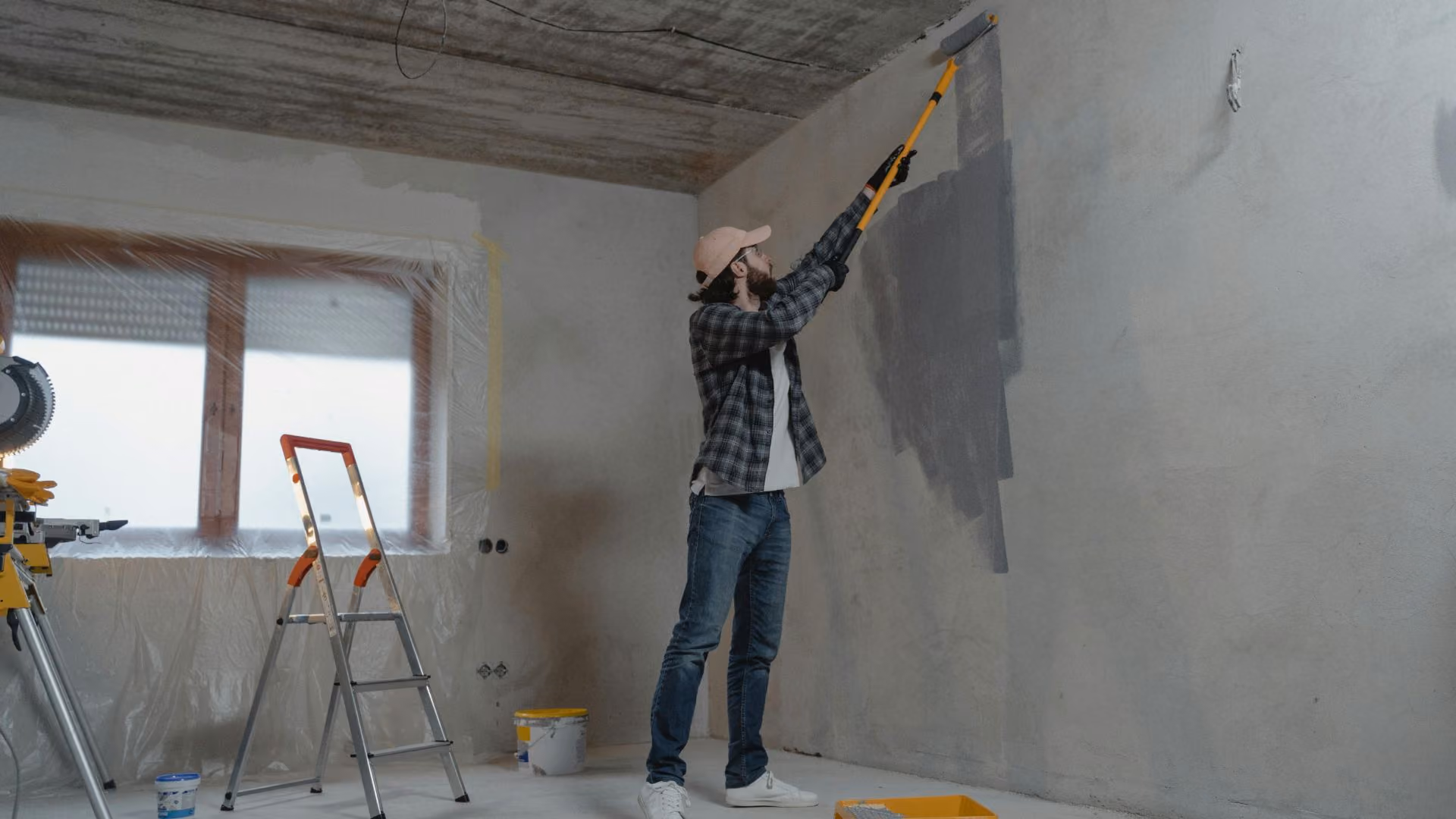 Person painting the wall of an apartment during a renovation project, with a ladder and covered windows in the background.