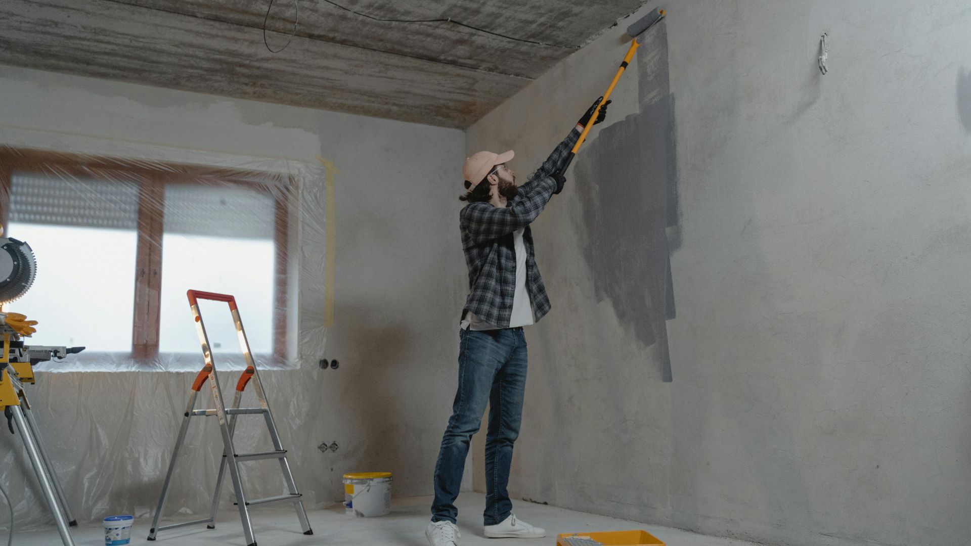Person painting the wall of an apartment during a renovation project, with a ladder and covered windows in the background.