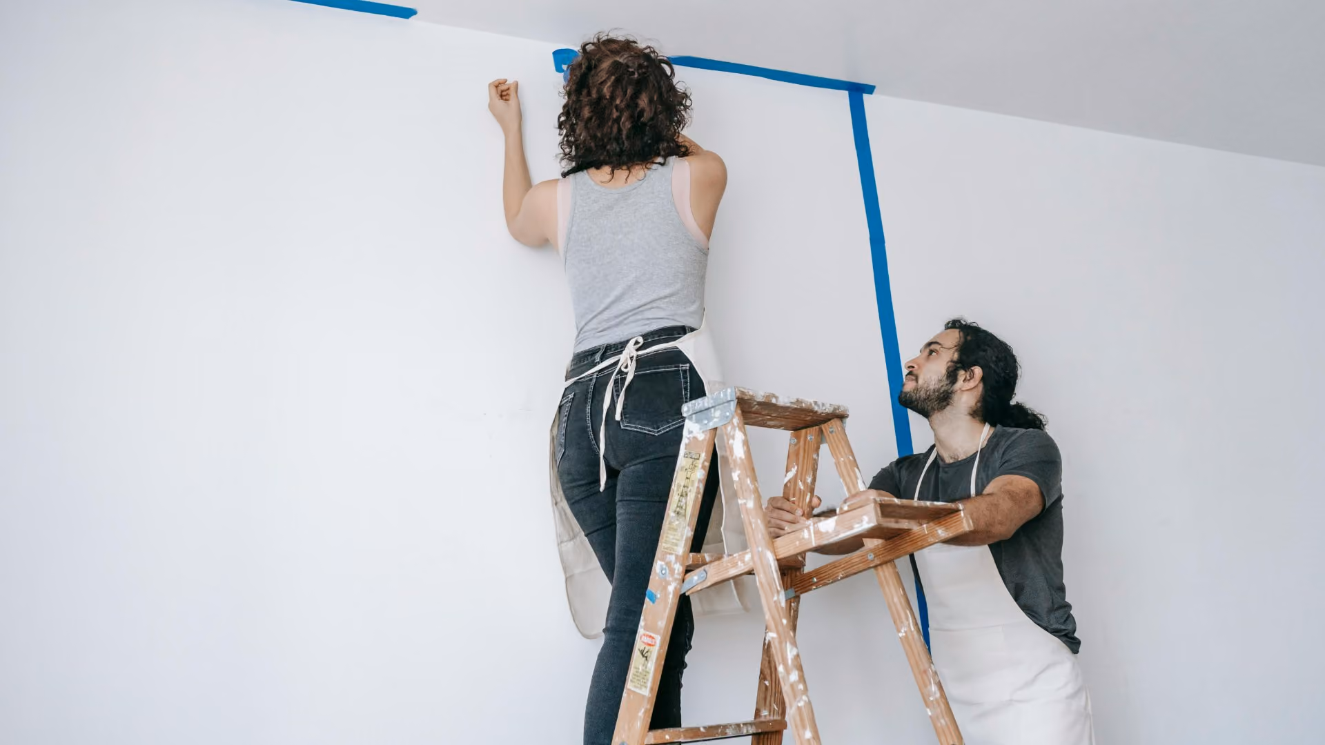 Two people preparing an interior wall for painting using painter's tape and a ladder.