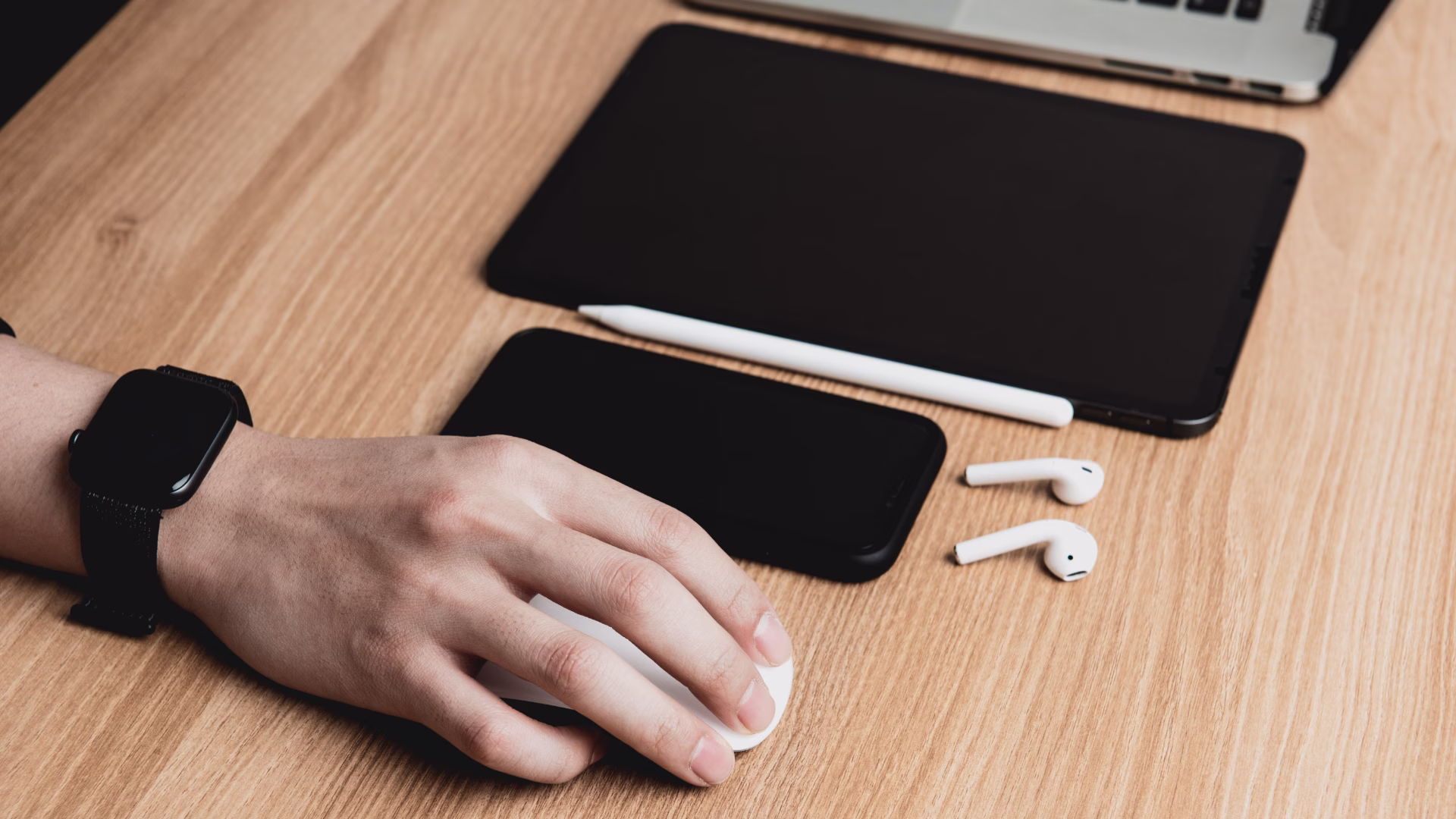 a person sitting at a desk with a smart phone, AirPods, laptop, mouse, and tablet