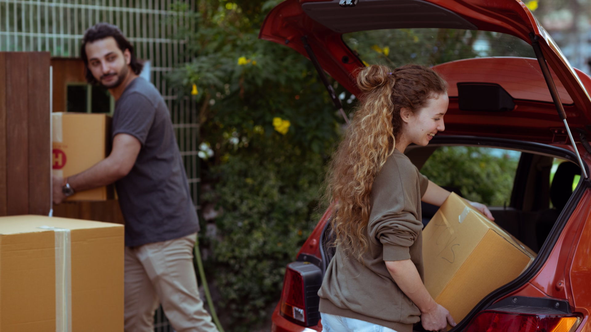 2 people unloading boxes from car trunk into a house