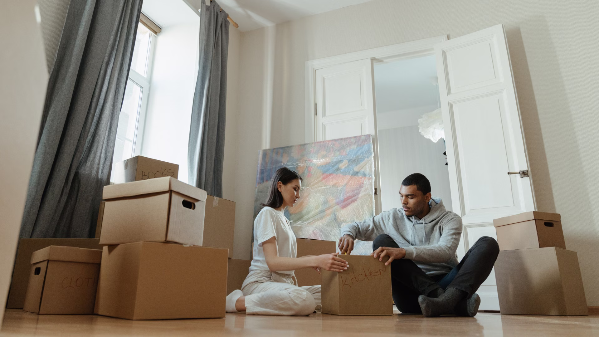 A bright, sunlit apartment living room filled with a few open moving boxes. A young couple is taking a break from unpacking