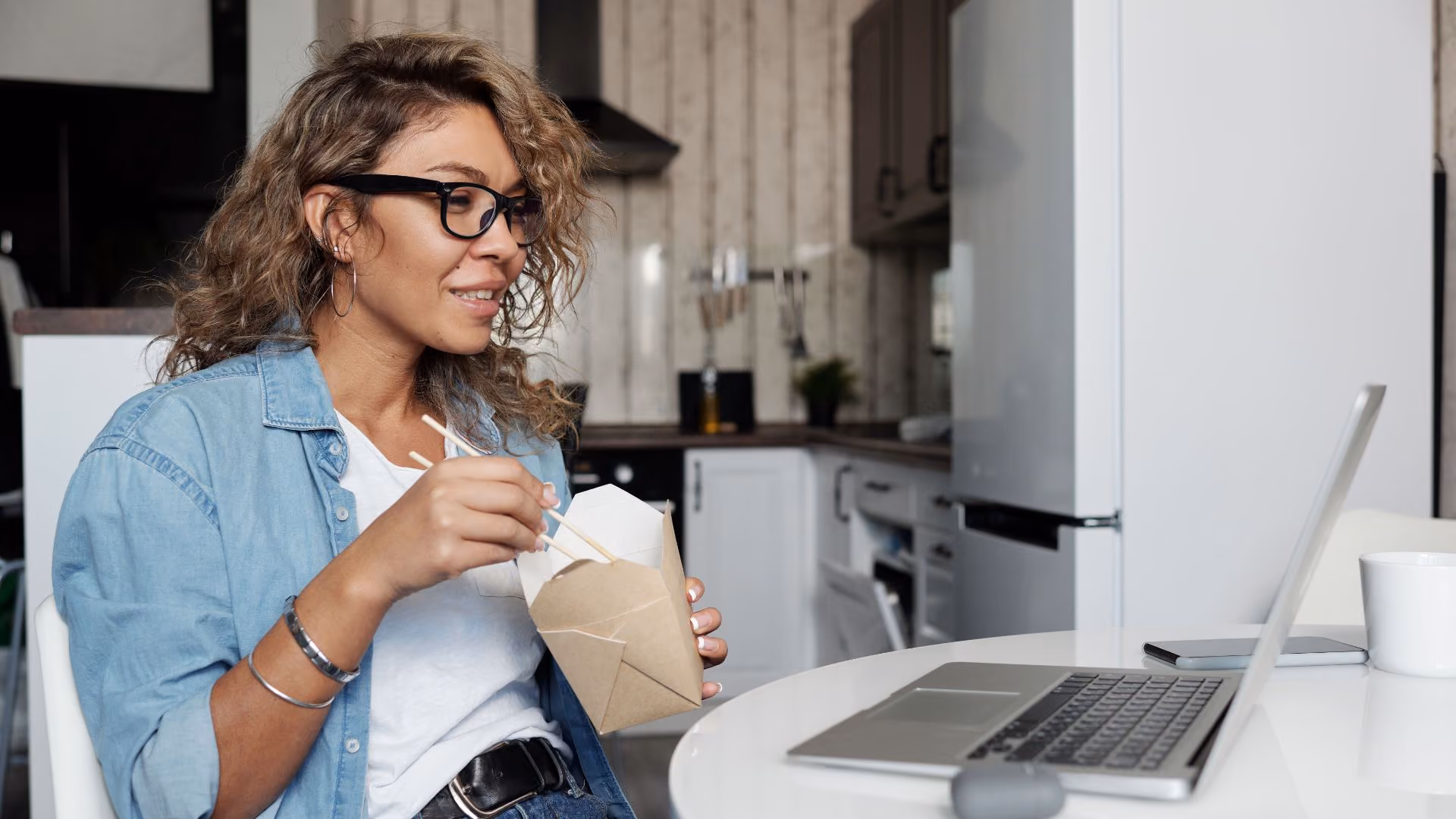 Person eating takeout while using a laptop in a modern apartment kitchen.