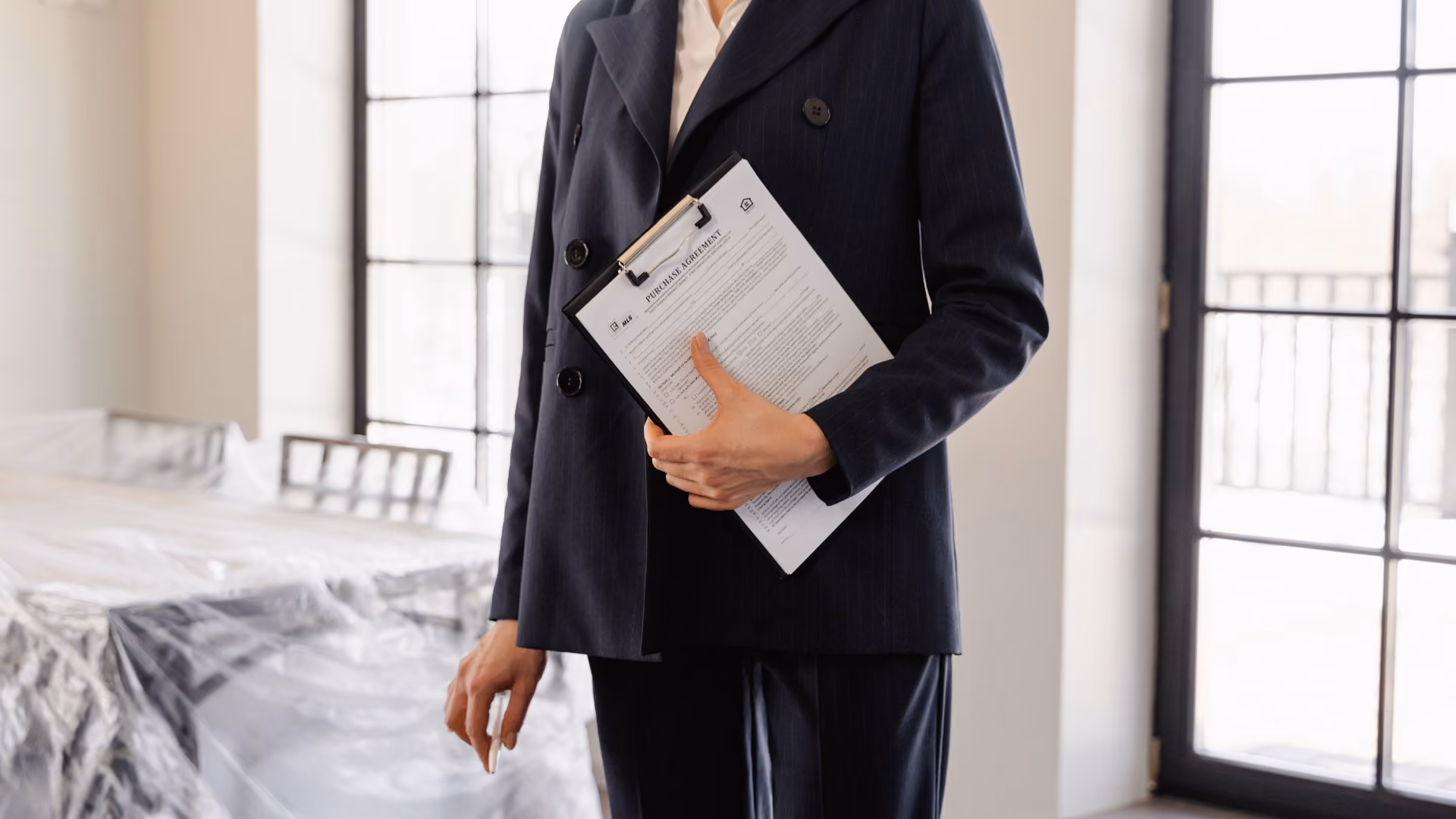 Person holding a clipboard with a purchase agreement while standing in an empty apartment with furniture covered in plastic wrap.