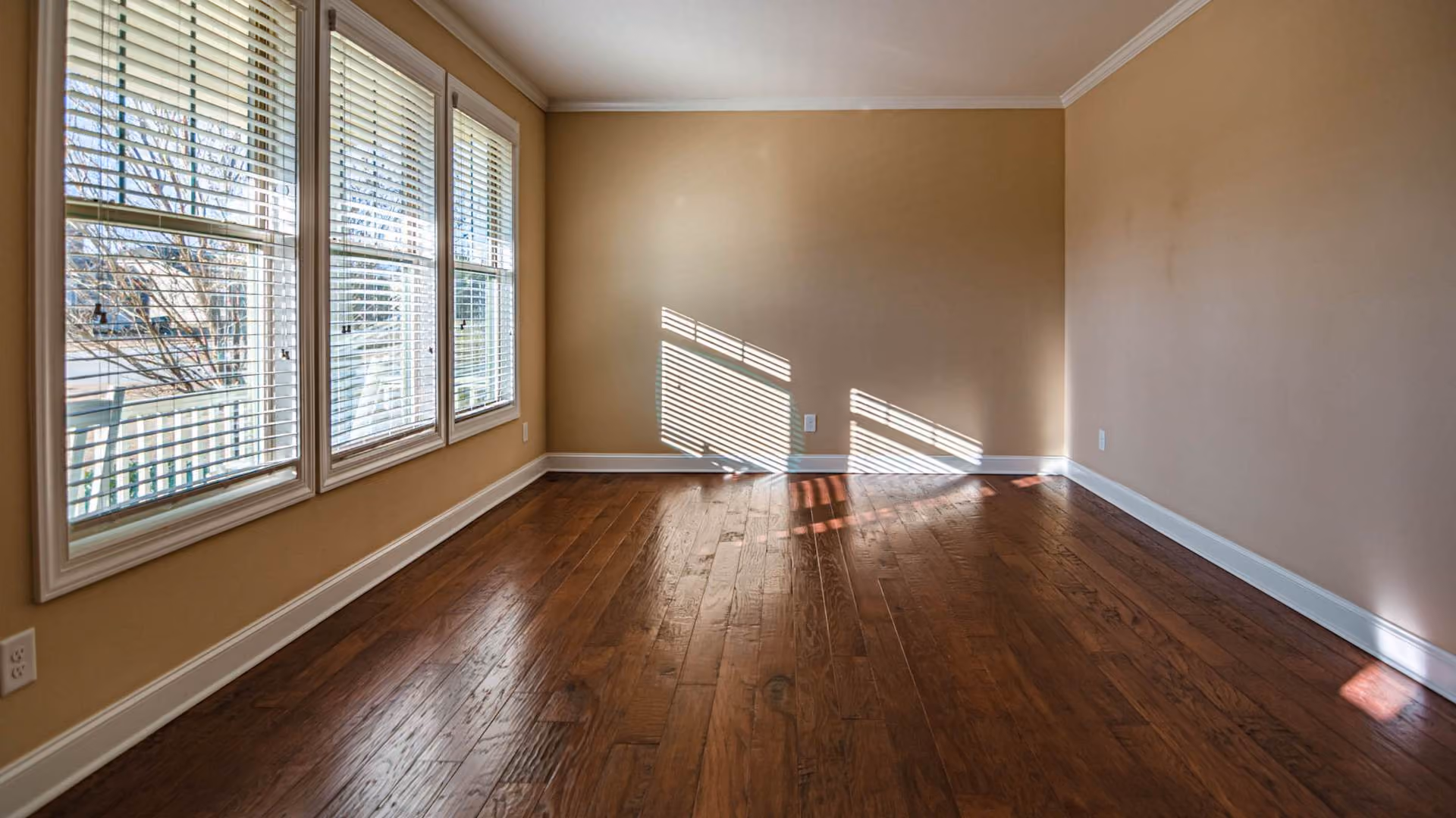 Empty apartment room with hardwood floors and natural light coming through window blinds.