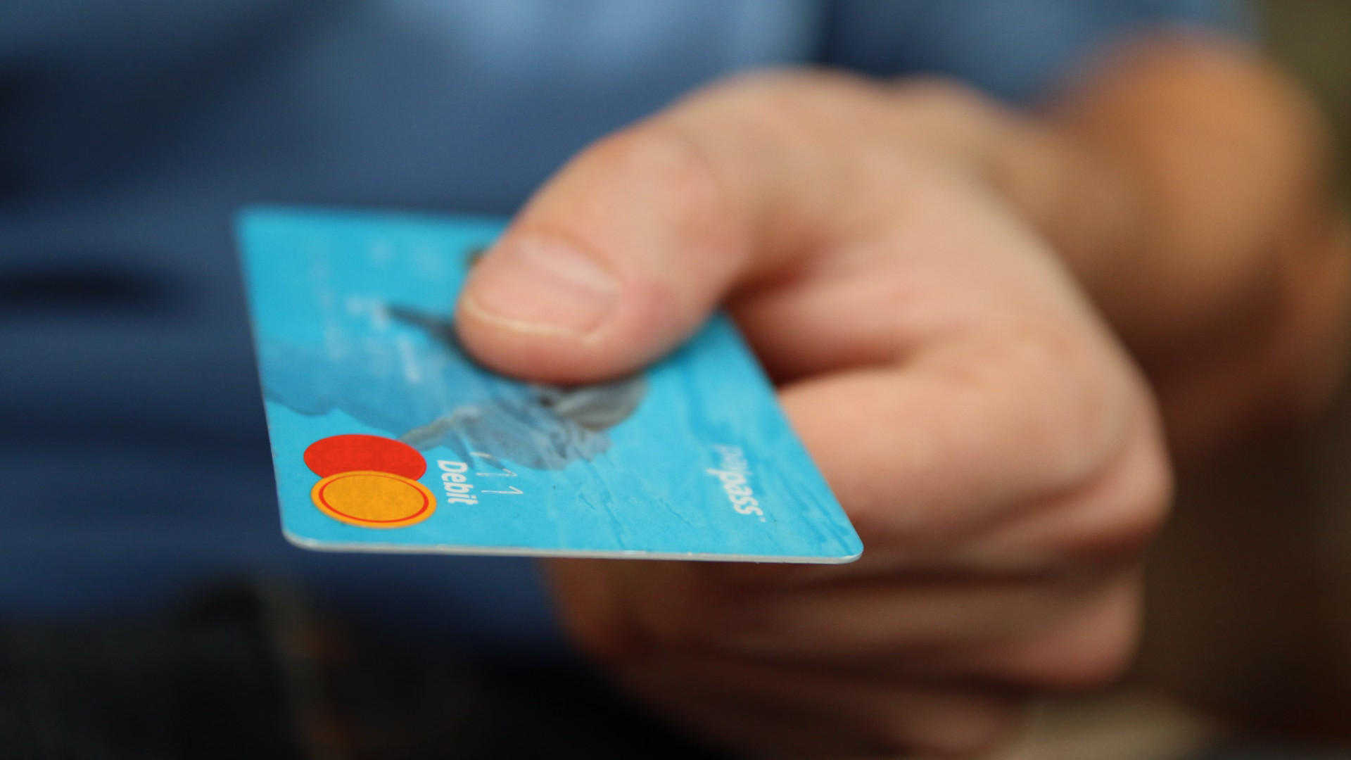 Person holding a credit card toward a silver laptop while working at a light wood desk