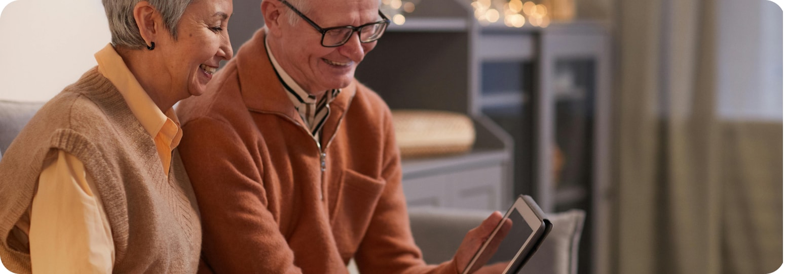 Older couple sitting together on a couch, smiling while looking at a tablet