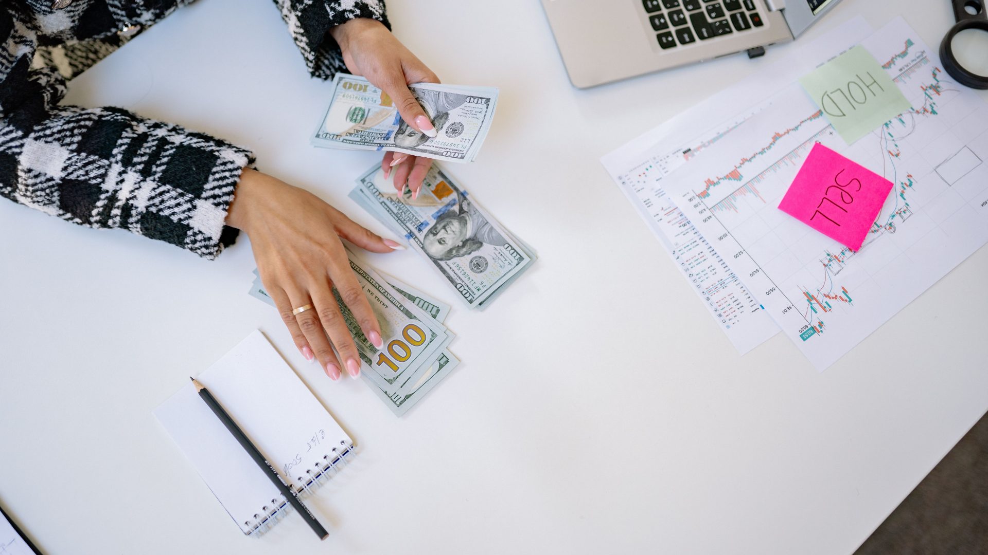 Person counting U.S. dollar bills at a desk beside printed stock market charts and a laptop.