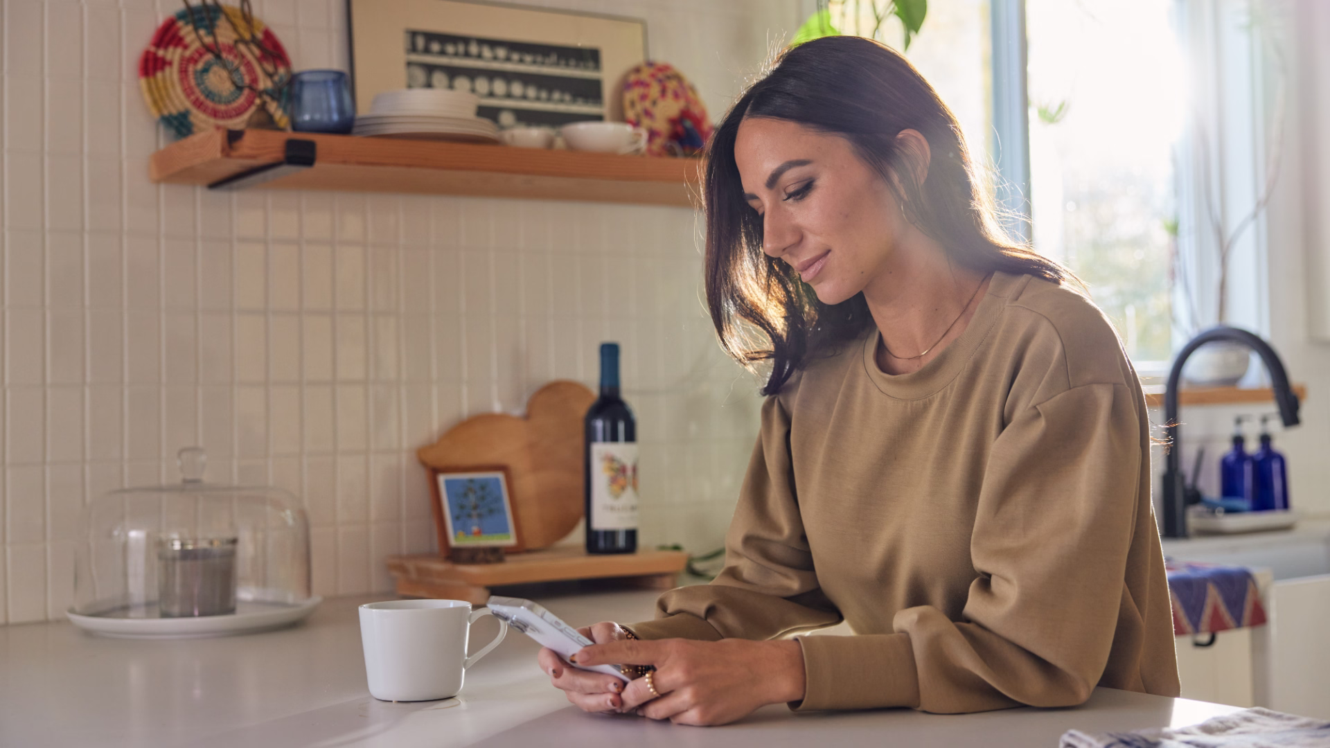 Woman in a warm kitchen checking her phone beside a mug and trailing pothos plants
