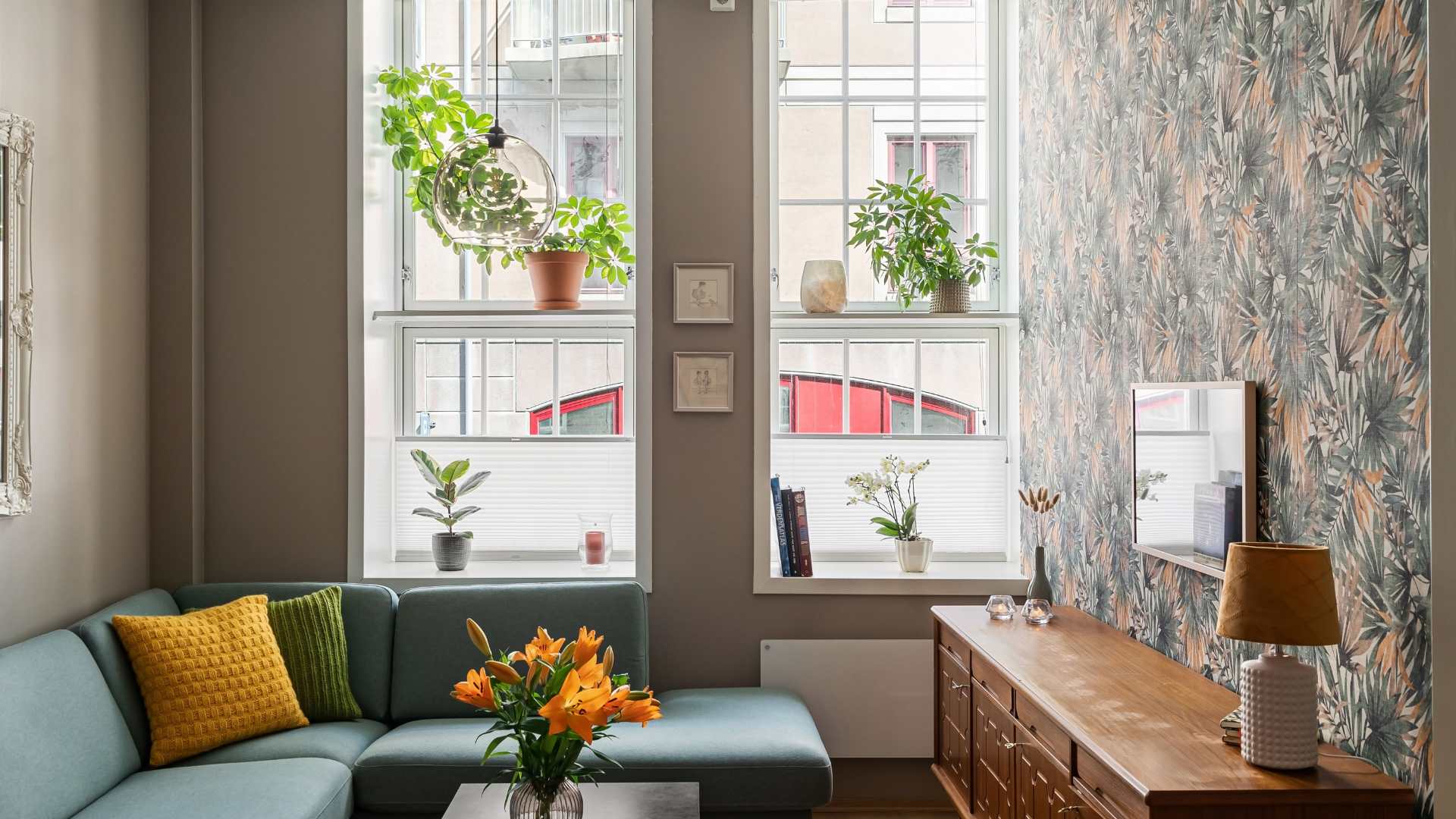 Cozy apartment living room with a teal sofa, patterned wallpaper, and plants near sunlit windows