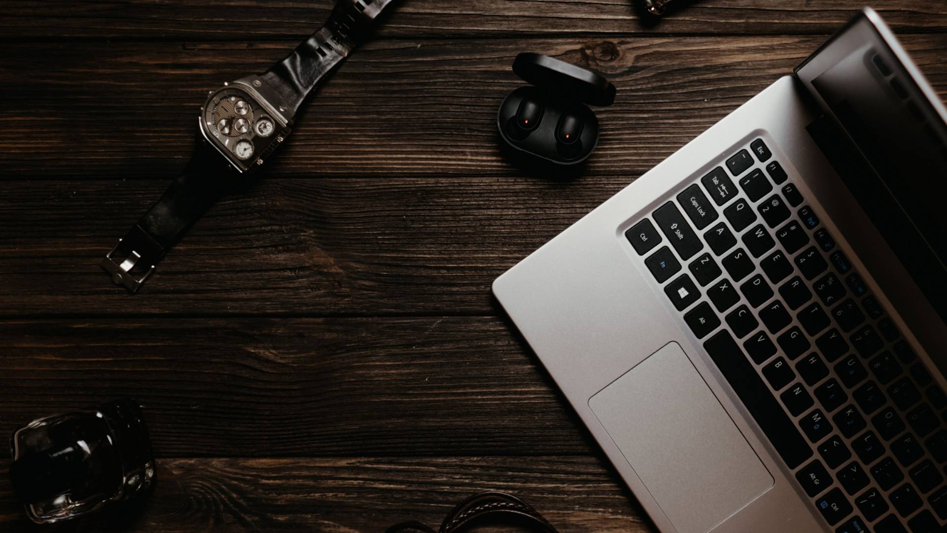 Flat lay of a watch, wireless earbuds, and laptop on a wooden desk