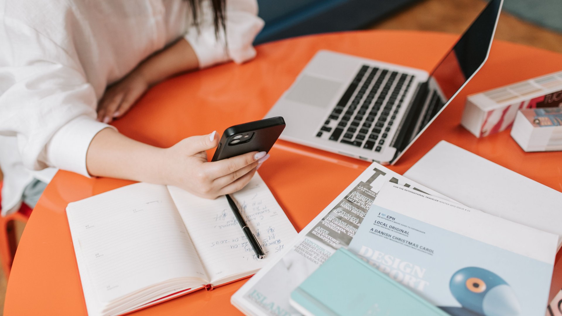 Person checking their phone next to a laptop, planner, and magazines at a table