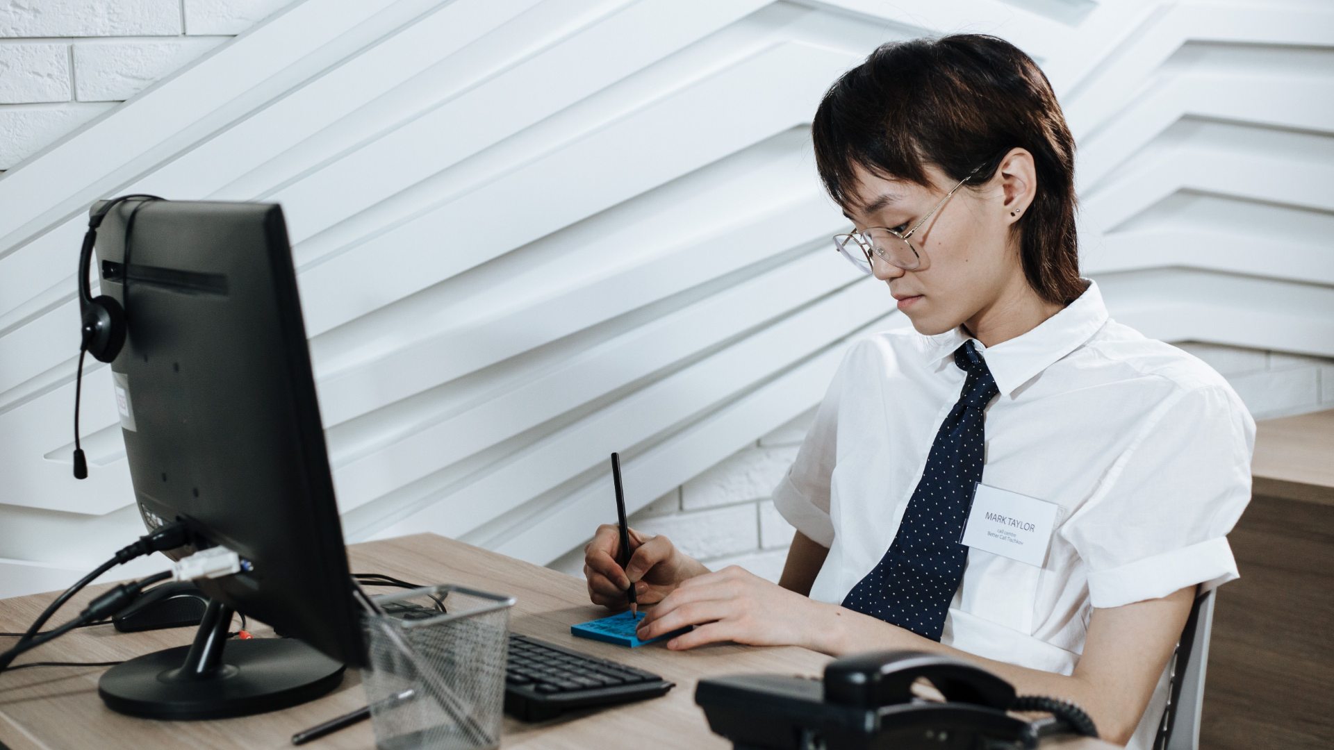 Person wearing glasses and a tie sitting at a desk with a computer monitor, writing on a notepad