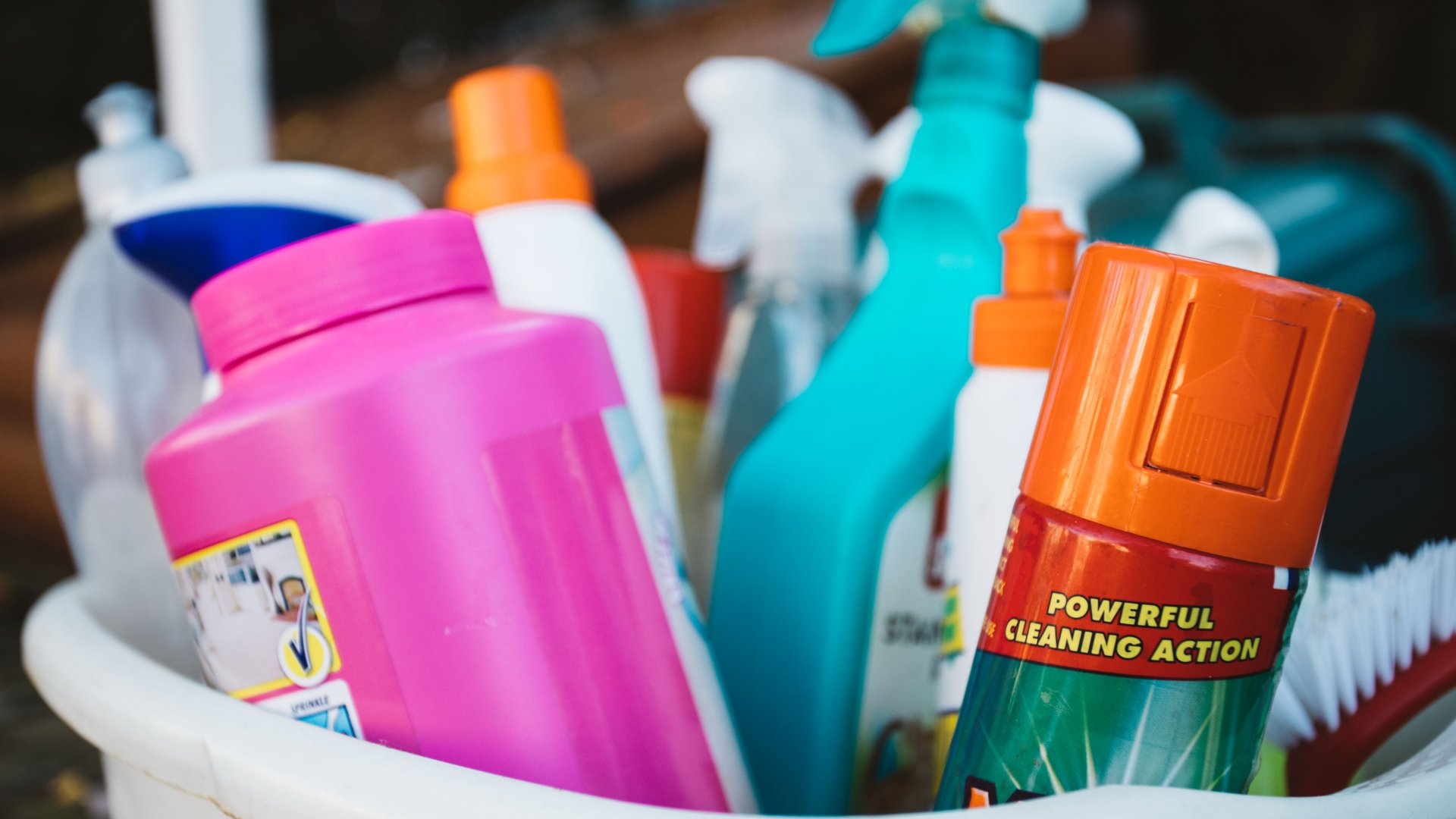 Cleaning bucket filled with cloths, spray bottles, and a bright green scrub brush