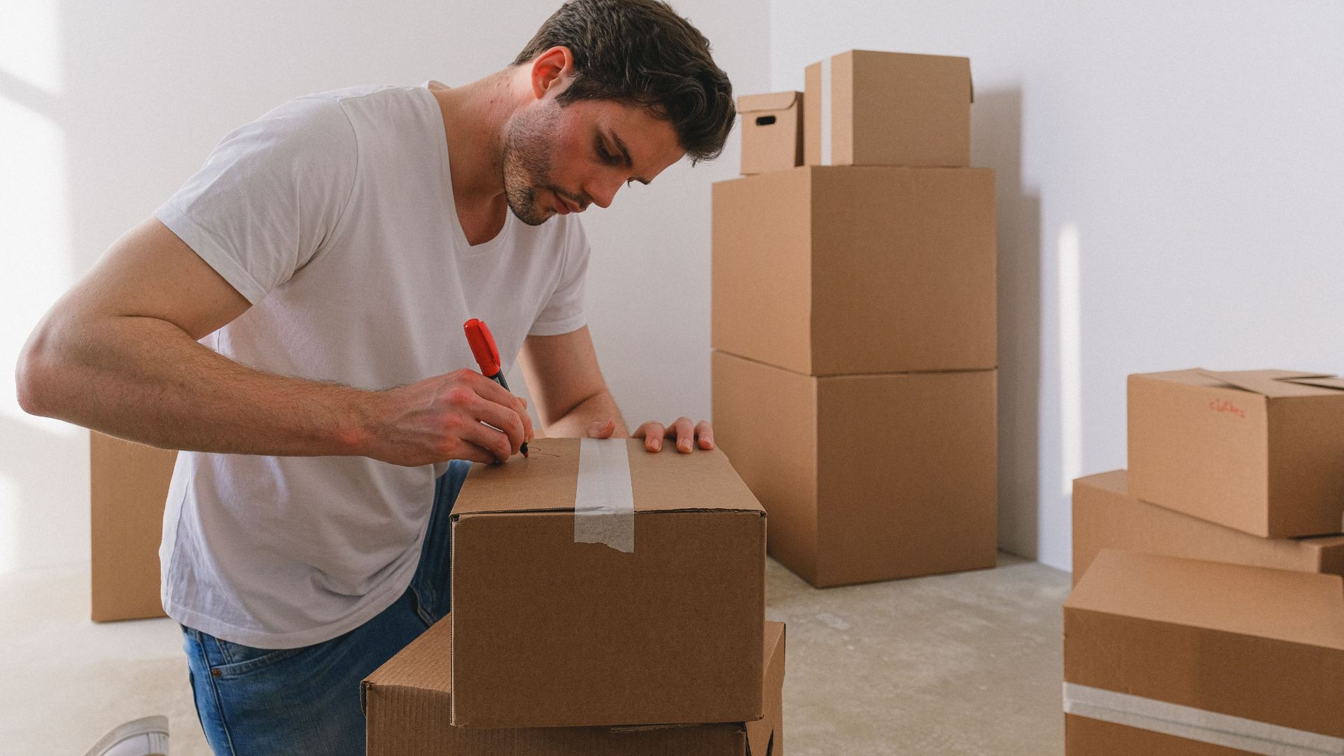 A man labeling a cardboard box with a red marker while packing up his apartment for a move.
