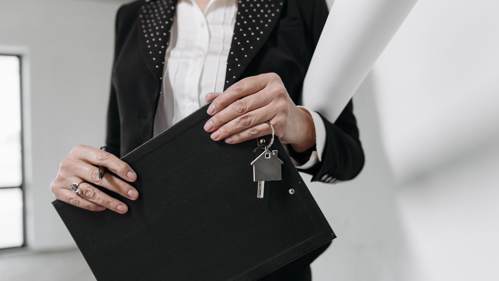 A person in a blazer holding a black portfolio folder with a house-shaped keychain and keys resting on top.